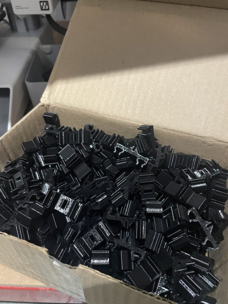 Caixa de papelão cheia de peças técnicas pretas idênticas, produzidas por impressão 3D na Nutss 3D para estoque digital industrial.